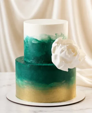 Bolo de dois andares com pintura aquarelada em tons de esmeralda e dourado, decorado com uma grande flor branca feita à mão. Peso do bolo a partir de 3,5 kg.